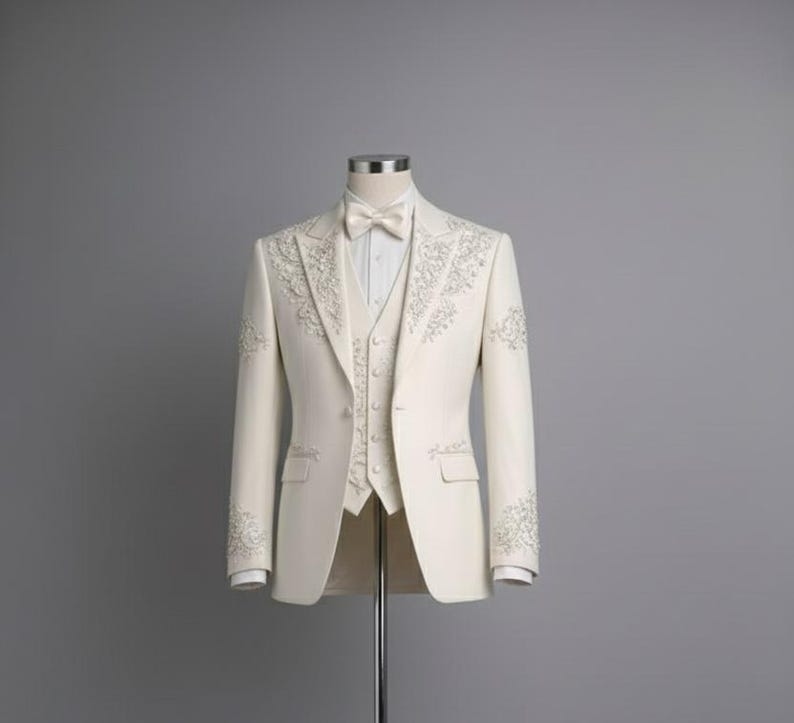 May include: Ivory three-piece suit with intricate floral embroidery. The suit includes a blazer, vest, and trousers, all in a solid ivory color. The blazer features a notched lapel and a bow tie. The suit is displayed on a mannequin against a gray background.
