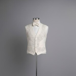 May include: Ivory vest and bow tie set on a mannequin. The vest features an intricate embroidered design in a lighter shade, with a matching bow tie. The ensemble is displayed against a gray backdrop, showcasing formal wear.