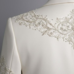 May include: Ivory-colored formal jacket with intricate beaded and embroidered embellishments. The design features delicate floral patterns and sparkling accents, creating an elegant and luxurious look. The jacket is tailored with a classic silhouette.