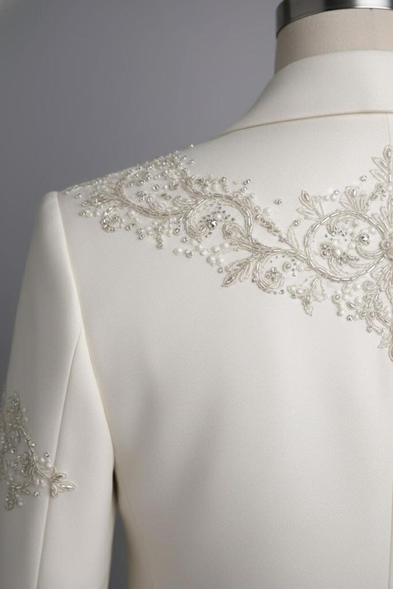 May include: Ivory-colored formal jacket with intricate beaded and embroidered embellishments. The design features delicate floral patterns and sparkling accents, creating an elegant and luxurious look. The jacket is tailored with a classic silhouette.