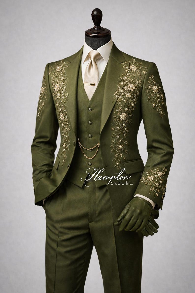 May include: Olive green three-piece suit with floral embroidery. The suit includes a jacket, vest, and trousers. The jacket features detailed floral embroidery with pearl accents. A gold chain and tie clip add a touch of elegance. The suit is displayed on a mannequin.