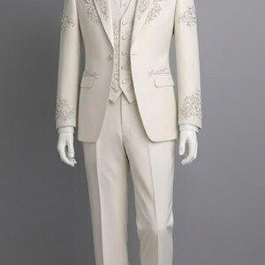 May include: Ivory three-piece suit on a mannequin. The suit features intricate floral embroidery on the lapels, cuffs, and vest. Includes a matching bow tie. The suit is tailored with a classic fit and is displayed against a gray backdrop.