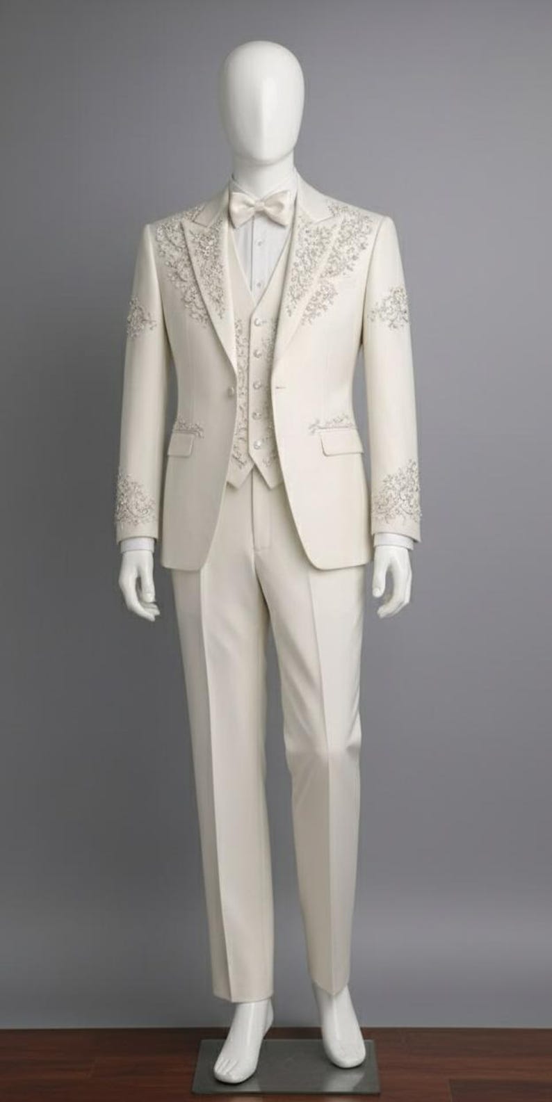 May include: Ivory three-piece suit on a mannequin. The suit features intricate floral embroidery on the lapels, cuffs, and vest. Includes a matching bow tie. The suit is tailored with a classic fit and is displayed against a gray backdrop.