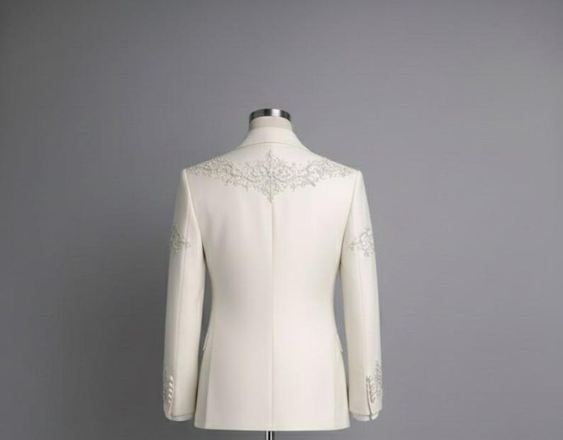 May include: Ivory-colored blazer with intricate silver embroidery on the shoulders and sleeves. The tailored jacket features a classic design with a two-button closure and a notched lapel. The back view showcases the detailed craftsmanship of the formal wear.