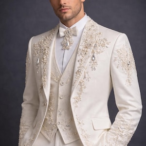 Op de afbeelding: Ivoor driedelig pak met ingewikkelde parel- en kristallen versieringen. Het pak bestaat uit een jasje, vest en broek. Het jasje en vest zijn versierd met gedetailleerde bloempatronen. Een witte vlinderdas en overhemd maken het formele ensemble compleet.