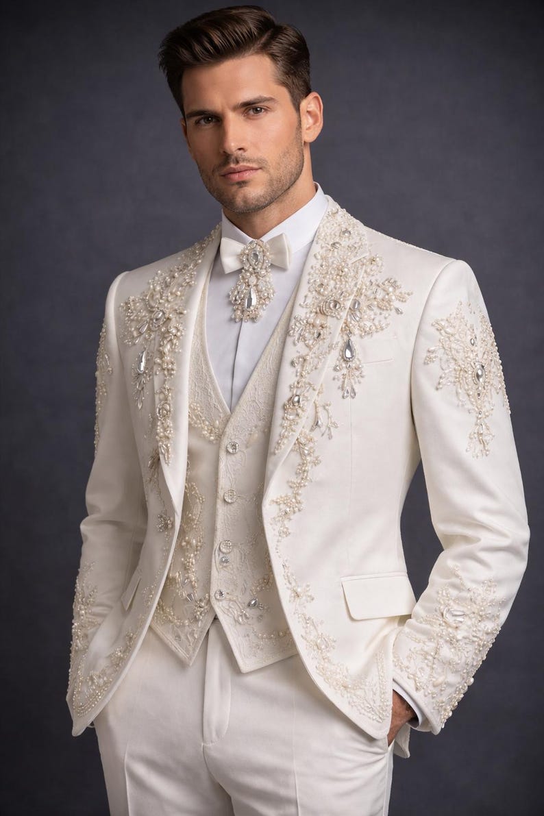 Op de afbeelding: Ivoor driedelig pak met ingewikkelde parel- en kristallen versieringen. Het pak bestaat uit een jasje, vest en broek. Het jasje en vest zijn versierd met gedetailleerde bloempatronen. Een witte vlinderdas en overhemd maken het formele ensemble compleet.