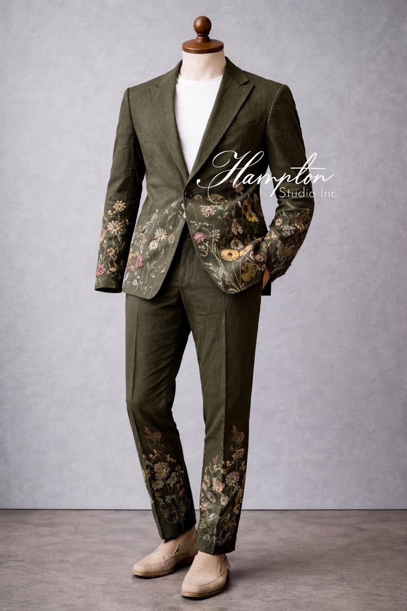 May include: Olive green suit with floral embroidery on the sleeves and pant legs. The suit includes a blazer and matching pants. The suit is displayed on a mannequin. The text "Hampton Studio Inc." is visible on the suit.
