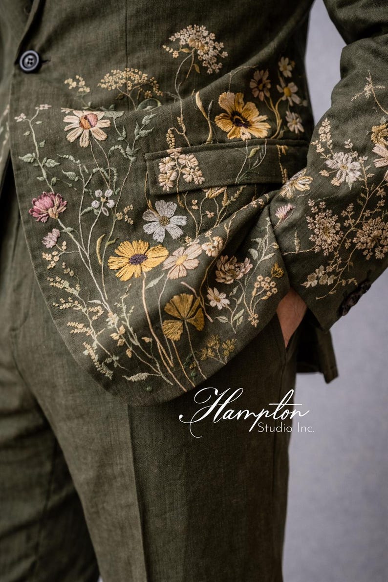 May include: Olive green blazer and pants set with intricate floral embroidery in yellow, white, and pink. The jacket features a notched lapel and a single-button closure. The text "Hampton Studio Inc." is visible.