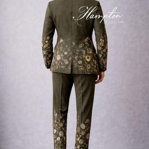 May include: Olive green suit with floral embroidery. The suit features a jacket and matching pants, both adorned with delicate flower patterns in shades of yellow and white. The text "Hampton Studio Inc." is on the back of the jacket.
