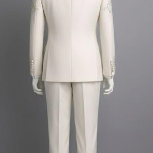 May include: Ivory-colored suit with intricate silver embroidery on the shoulders and sleeves. The suit includes a tailored jacket and matching trousers, displayed on a mannequin. A formal wear ensemble.
