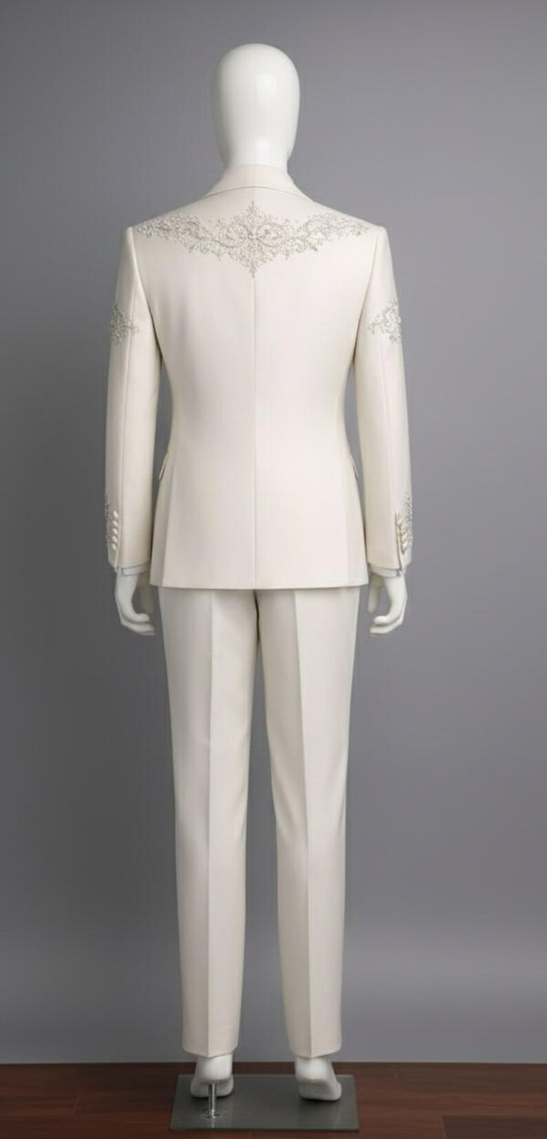 May include: Ivory-colored suit with intricate silver embroidery on the shoulders and sleeves. The suit includes a tailored jacket and matching trousers, displayed on a mannequin. A formal wear ensemble.