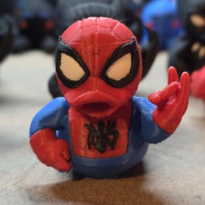 K&ouml;nnte beinhalten: Eine 3D-gedruckte Spider-Man-Figur im Enten-Design. Sie hat einen roten Kopf und H&auml;nde, einen blauen K&ouml;rper und wei&szlig;e Augen. Ein schwarzes Spinnenemblem befindet sich auf der Brust. Die Figur ist mit erhobener Hand dargestellt.