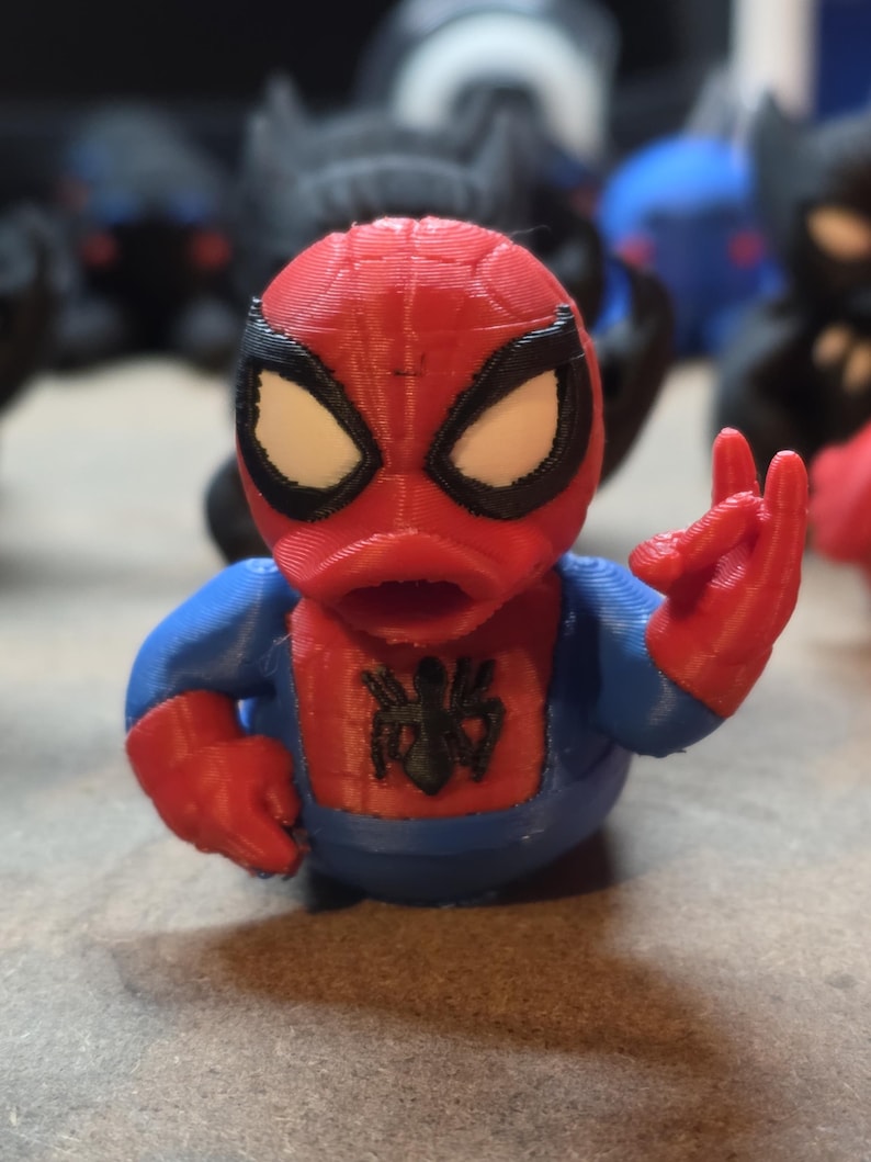 K&ouml;nnte beinhalten: Eine 3D-gedruckte Spider-Man-Figur im Enten-Design. Sie hat einen roten Kopf und H&auml;nde, einen blauen K&ouml;rper und wei&szlig;e Augen. Ein schwarzes Spinnenemblem befindet sich auf der Brust. Die Figur ist mit erhobener Hand dargestellt.