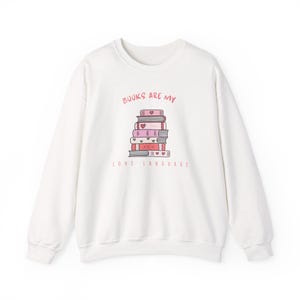 Könnte beinhalten: Weißes Sweatshirt mit dem Text "BOOKS ARE MY LOVE LANGUAGE" in Rosa. Die Grafik zeigt einen Stapel Bücher mit Herzen und dekorativen Elementen. Ein gemütliches, lässiges Oberteil für Bücherliebhaber.