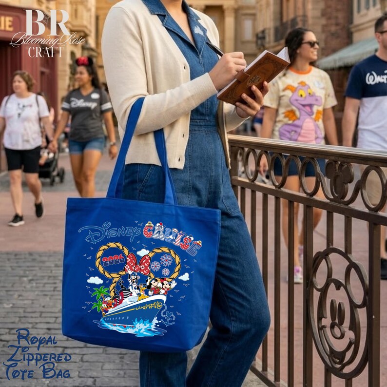 May include: A royal blue tote bag featuring a graphic of a cruise ship with Mickey and Minnie Mouse, rope-style ears, the text Disney Cruise 2026, and small fireworks. The bag has a zipper closure and is held by a person wearing a cream cardigan.