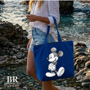 May include: A blue tote bag featuring a smiling Mickey Mouse graphic. The bag has a zipper closure and a rectangular shape. The bag is held by a person near the ocean. The Blooming Rose Craft logo is visible in the bottom left corner.