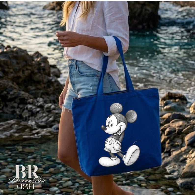 May include: A blue tote bag featuring a smiling Mickey Mouse graphic. The bag has a zipper closure and a rectangular shape. The bag is held by a person near the ocean. The Blooming Rose Craft logo is visible in the bottom left corner.