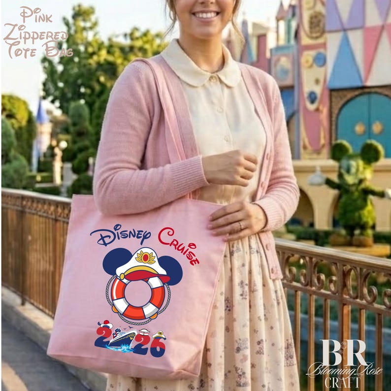 Puede incluir: Un bolso tote de color rosa claro con cremallera que presenta un gr&aacute;fico de un salvavidas rojo y blanco con orejas de Mickey Mouse y una gorra de capit&aacute;n. El texto Disney Cruise aparece sobre el gr&aacute;fico y 2026 debajo con un peque&ntilde;o crucero.