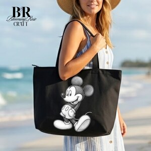 May include: Black tote bag featuring a black and white illustration of Mickey Mouse. The bag has a zipper closure and black handles. The text "BR Blooming Rose Craft" is in the upper left corner. The bag is being held on a beach.