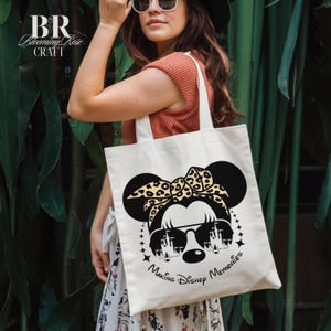May include: Cream-colored tote bag featuring a black graphic of a mouse with sunglasses and a leopard print bow. The text "Making Disney Memories" is below the graphic. The bag has white handles and the brand name "Blooming Rose Craft" is at the top left.