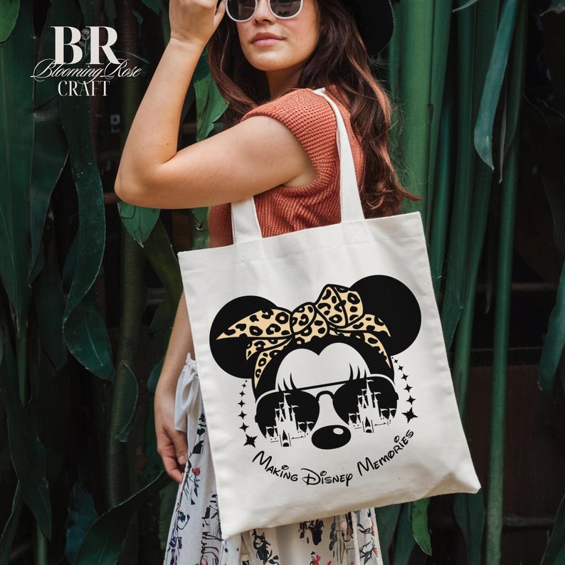 May include: Cream-colored tote bag featuring a black graphic of a mouse with sunglasses and a leopard print bow. The text "Making Disney Memories" is below the graphic. The bag has white handles and the brand name "Blooming Rose Craft" is at the top left.