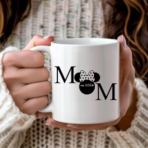 May include: White ceramic mug with the word "MOM" in black, featuring a Minnie Mouse bow and silhouette. The mug also includes "est 2024" in a circular design. The mug is held by a person wearing a white sweater.