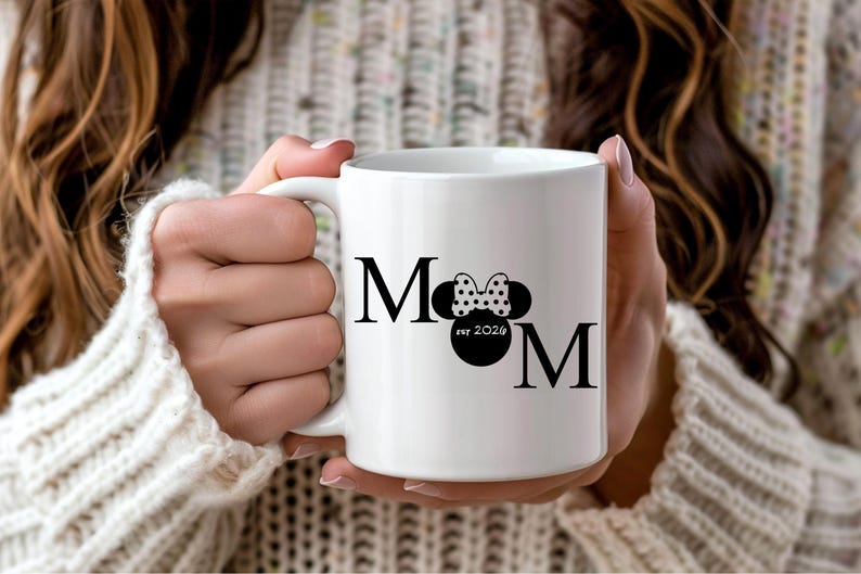 May include: White ceramic mug with the word "MOM" in black, featuring a Minnie Mouse bow and silhouette. The mug also includes "est 2024" in a circular design. The mug is held by a person wearing a white sweater.