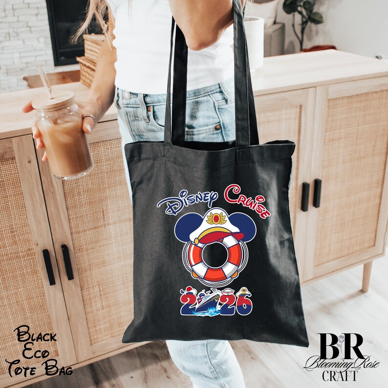 Puede incluir: Un bolso tote negro con un gr&aacute;fico de un salvavidas que lleva una gorra de capit&aacute;n con orejas de Mickey Mouse. Sobre el gr&aacute;fico aparece el texto Disney Cruise y debajo el n&uacute;mero 2026 con un crucero ilustrado entre los n&uacute;meros.