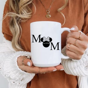 May include: White ceramic mug with the word "Mom" in black, featuring a Minnie Mouse bow and the text "est 2024". The mug has a large handle and is held by a person wearing a white sweater.