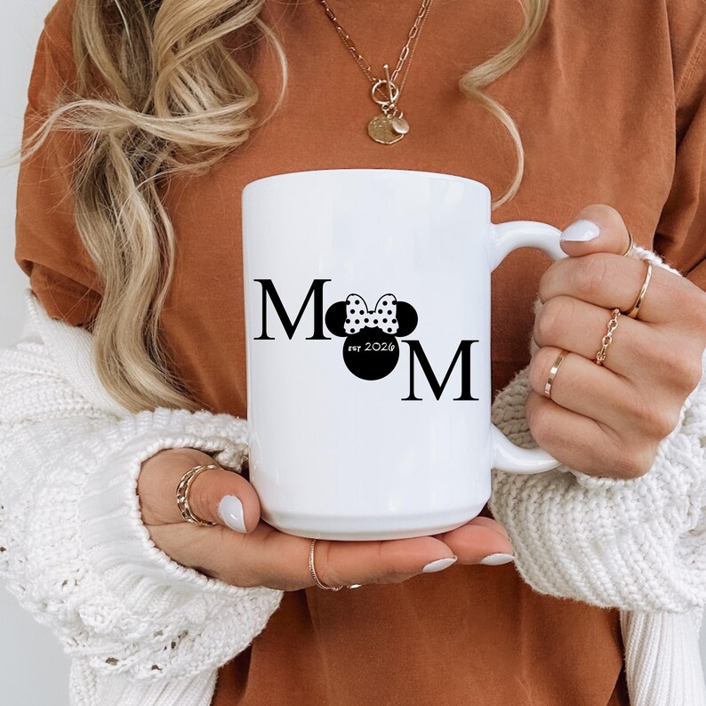 May include: White ceramic mug with the word "Mom" in black, featuring a Minnie Mouse bow and the text "est 2024". The mug has a large handle and is held by a person wearing a white sweater.