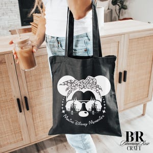 May include: Black tote bag featuring a white graphic of a mouse with sunglasses, a bow, and a castle silhouette. The text "Making Disney Memories" is below the graphic. The bag is held by a person.