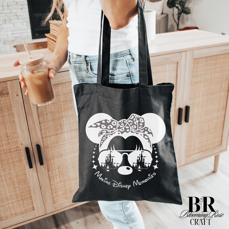 May include: Black tote bag featuring a white graphic of a mouse with sunglasses, a bow, and a castle silhouette. The text "Making Disney Memories" is below the graphic. The bag is held by a person.