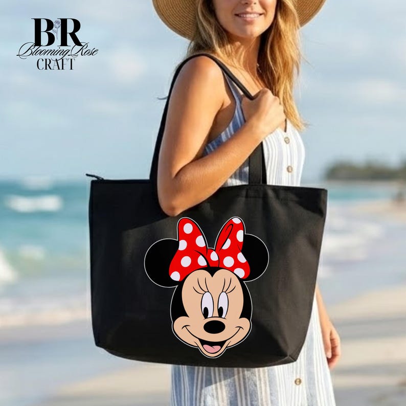 Puede incluir: Bolso tote negro con un gr&aacute;fico de Minnie Mouse con un lazo rojo con lunares blancos. El bolso tiene asas negras y cierre de cremallera. Una persona sostiene el bolso en una playa.