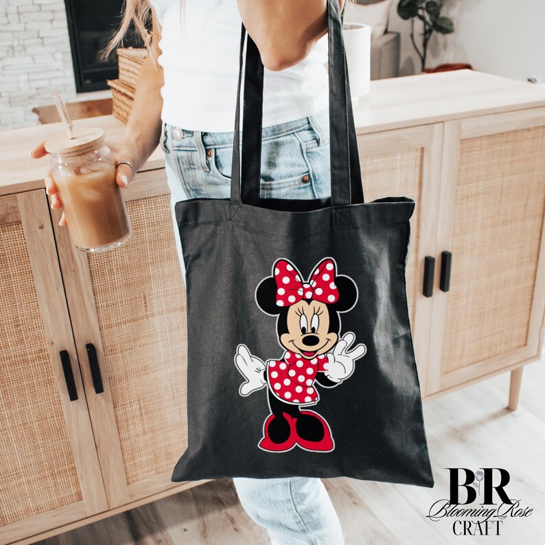 Peut inclure: Sac fourre-tout noir avec un motif Minnie Mouse. Le motif Minnie Mouse comprend une robe et un n&oelig;ud &agrave; pois rouges. Le sac est tenu par une personne portant un jean bleu. Les mots "BR Blooming Rose CRAFT" sont imprim&eacute;s en bas &agrave; droite.