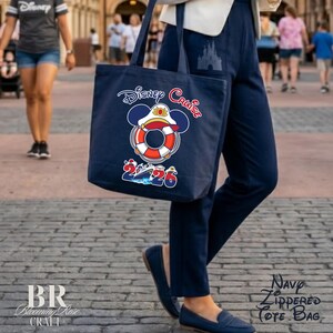 Puede incluir: Un bolso tote azul marino con un gr&aacute;fico de un salvavidas con orejas de Mickey Mouse y una gorra de capit&aacute;n. Debajo del gr&aacute;fico, el texto dice Disney Cruise 2026 con una peque&ntilde;a ilustraci&oacute;n de un crucero integrada en los n&uacute;meros.