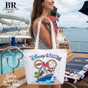 May include: A white tote bag featuring a graphic of a cruise ship with Disney characters, the text Disney Cruise, and the year 2026. The design includes rope-style Mickey Mouse ears, palm trees, and fireworks. The bag has two shoulder straps.