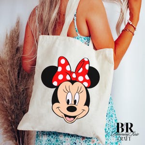 Puede incluir: Un bolso tote color crema con un gran gr&aacute;fico de la cara de Minnie Mouse. Minnie tiene una cara negra, un lazo rojo con lunares blancos. El bolso tiene dos asas color crema y el texto "BR Blooming Rose CRAFT" en la esquina inferior derecha.