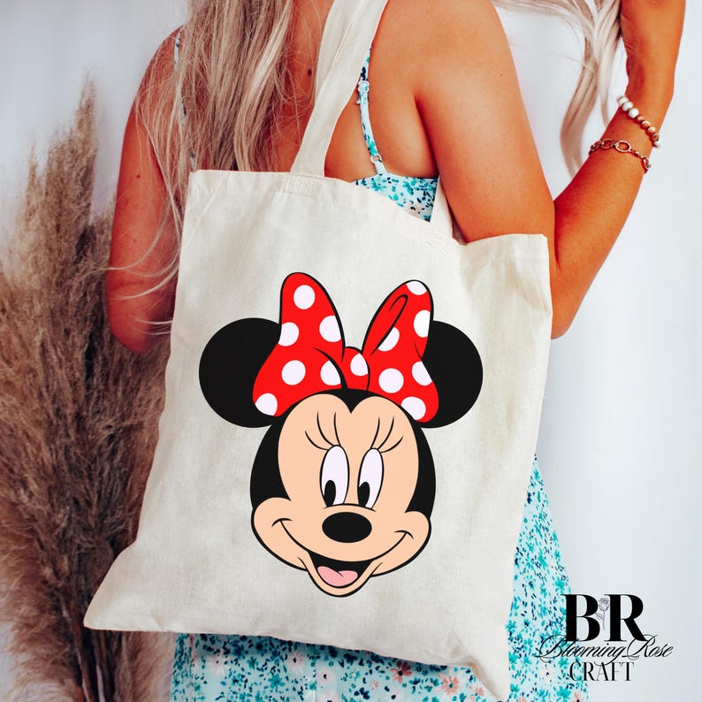 Puede incluir: Un bolso tote color crema con un gran gr&aacute;fico de la cara de Minnie Mouse. Minnie tiene una cara negra, un lazo rojo con lunares blancos. El bolso tiene dos asas color crema y el texto "BR Blooming Rose CRAFT" en la esquina inferior derecha.