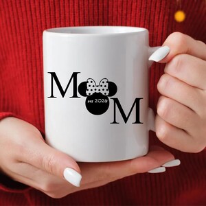 May include: White ceramic mug with the word "MOM" in black, featuring a Minnie Mouse-inspired design. The design includes a polka-dot bow and the text "est 2026". The mug is held by a person wearing a red sweater.