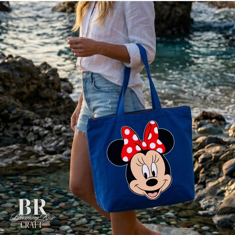 Puede incluir: Un bolso tote azul con un gr&aacute;fico de Minnie Mouse con un lazo rojo con lunares blancos. El bolso tiene cierre de cremallera y asas azules. El bolso lo lleva una persona en una playa.
