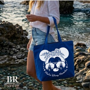 May include: A blue tote bag featuring a white graphic of a cartoon character with sunglasses and a bow. The text "Making Disney Memories" is below the graphic. The bag has two handles and a zipper closure.