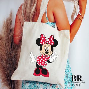 Peut inclure: Un sac fourre-tout cr&egrave;me avec un motif de Minnie Mouse. Minnie est repr&eacute;sent&eacute;e dans une robe et un n&oelig;ud &agrave; pois rouges, avec des oreilles et des chaussures noires. Le sac a deux anses et est port&eacute; par une personne.