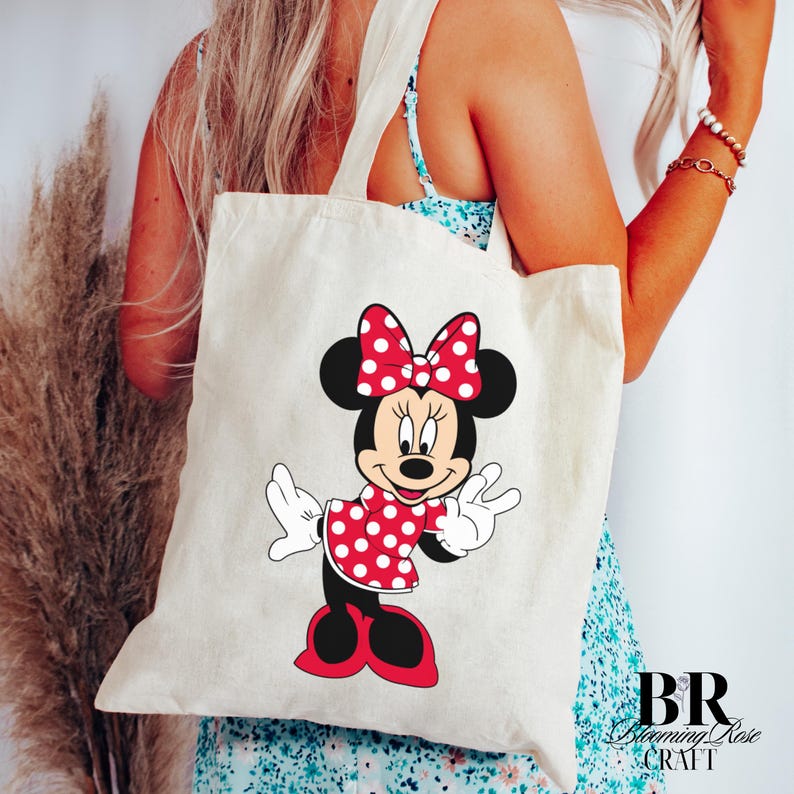 Peut inclure: Un sac fourre-tout cr&egrave;me avec un motif de Minnie Mouse. Minnie est repr&eacute;sent&eacute;e dans une robe et un n&oelig;ud &agrave; pois rouges, avec des oreilles et des chaussures noires. Le sac a deux anses et est port&eacute; par une personne.