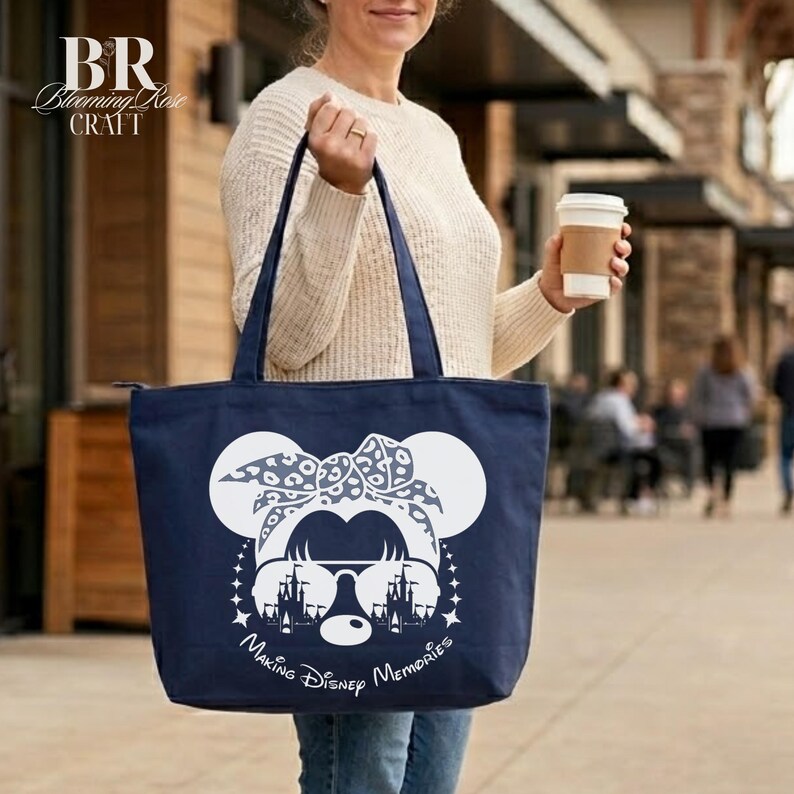 May include: Navy blue tote bag featuring a white Minnie Mouse design with sunglasses and a bow. The text "Making Disney Memories" is below the graphic. The bag has two handles and a zipper closure. The Blooming Rose Craft logo is in the upper left corner.