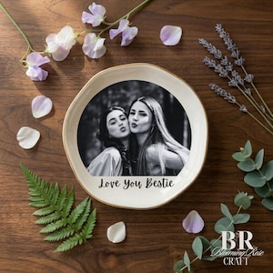 May include: A circular, cream-coloured ceramic plate featuring a black and white photograph of two women kissing. The plate has a gold rim and the words "Love You Bestie" are printed beneath the photo. The plate is surrounded by flowers and foliage.