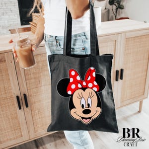 Puede incluir: Bolso tote negro con un dise&ntilde;o de Minnie Mouse, con un lazo rojo con lunares blancos. El bolso tiene asas negras y est&aacute; hecho de un material resistente. El logotipo de Blooming Rose Craft es visible en la esquina inferior derecha.