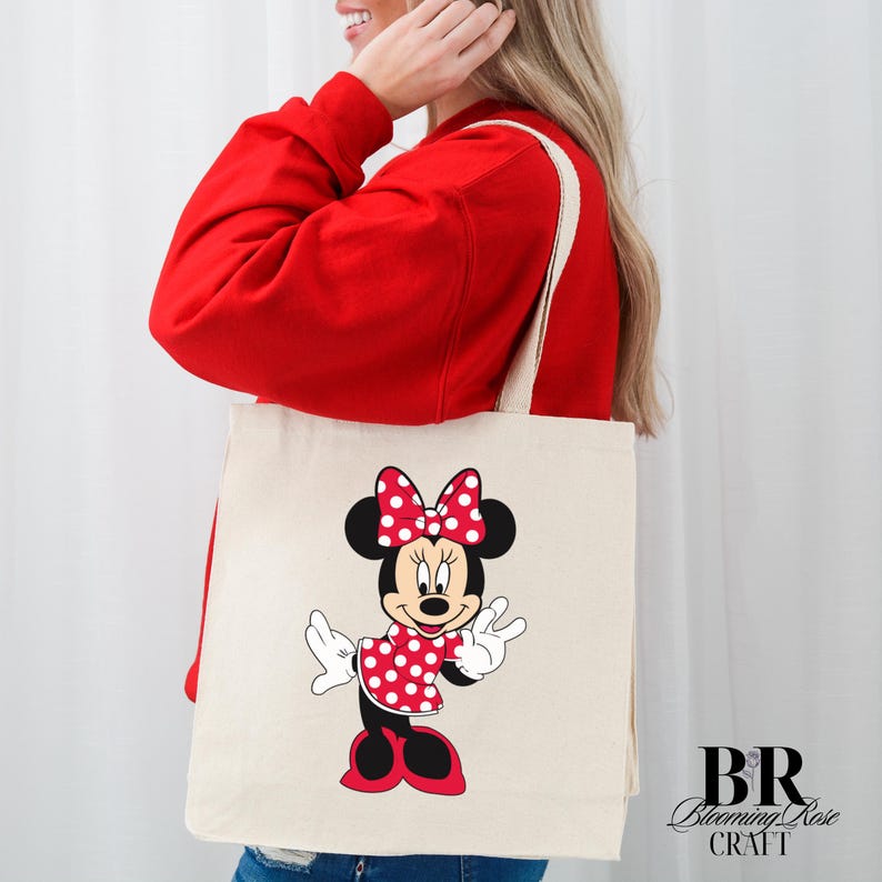 Peut inclure: Un sac fourre-tout cr&egrave;me avec un motif de Minnie Mouse. Minnie est repr&eacute;sent&eacute;e dans une robe et un n&oelig;ud &agrave; pois rouges, avec des oreilles et des chaussures noires. Le sac a des sangles cr&egrave;me et le texte "BR Blooming Rose CRAFT" en bas &agrave; droite.