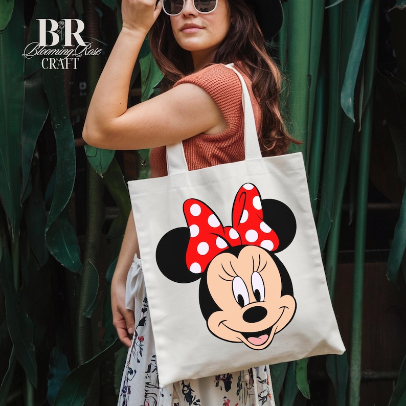 Puede incluir: Un bolso tote de color crema con un gr&aacute;fico de Minnie Mouse. Minnie Mouse tiene un lazo rojo con lunares blancos, orejas negras y una cara sonriente. La bolsa tiene el texto "BR Blooming Rose Craft" en la esquina superior izquierda.