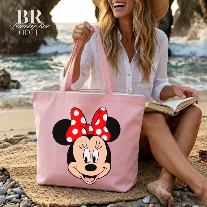 Puede incluir: Un bolso tote de color rosa claro con un dise&ntilde;o de Minnie Mouse, con un lazo rojo con lunares blancos. El bolso tiene asas rosas y el texto "BR Blooming Rose Craft". El bolso est&aacute; en una playa.