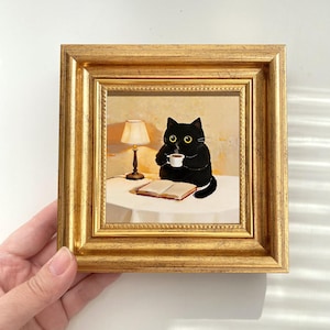 Puede incluir: Pintura enmarcada que representa un gato negro sentado en una mesa, sosteniendo una taza de café. Una pequeña lámpara ilumina la escena, y un libro abierto descansa sobre la mesa. El marco es dorado.
